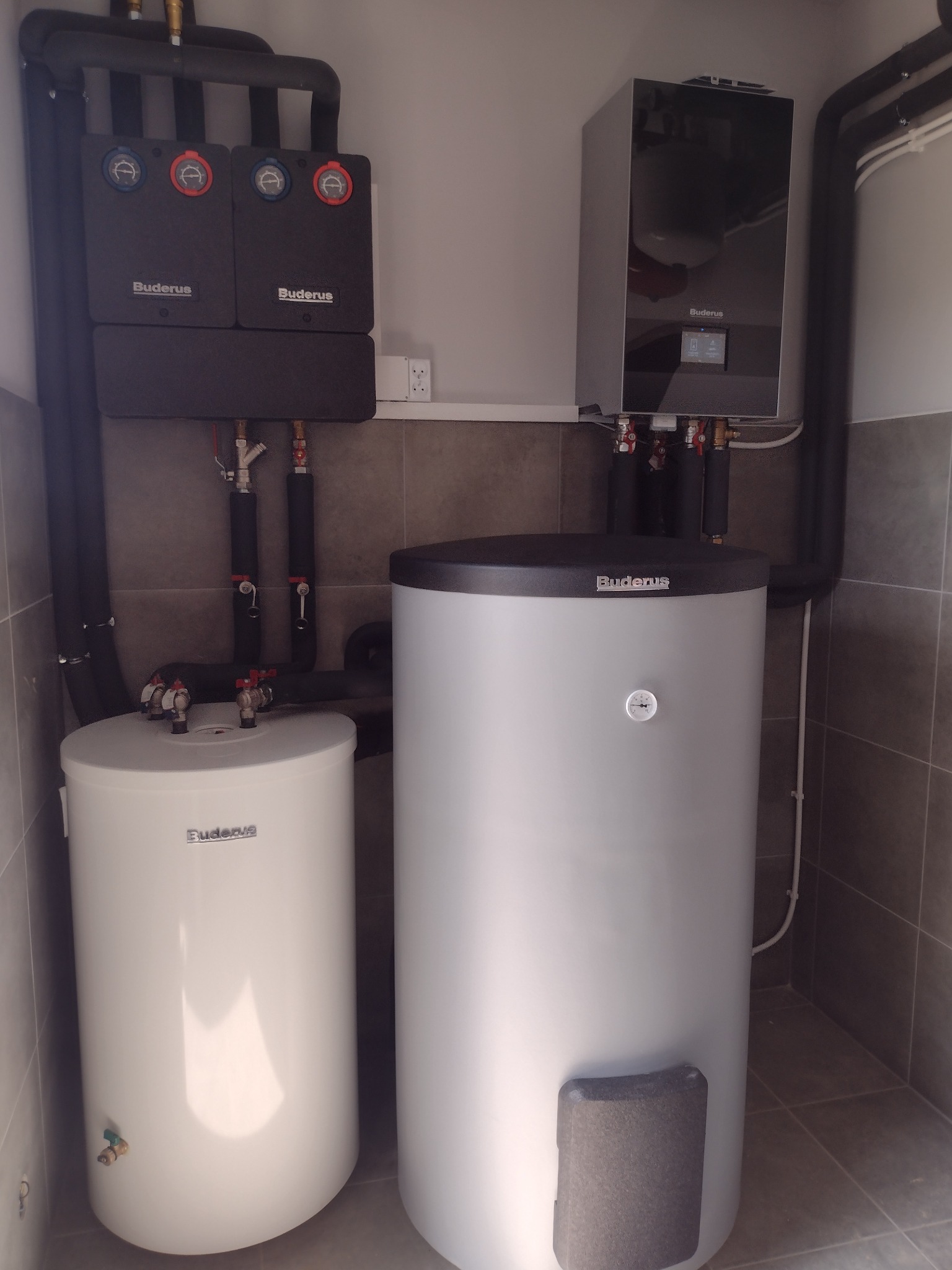 Nowoczesna kotłownia z dwoma zasobnikami ciepłej wody marki Buderus, czarnymi rurami w izolacji, zaworami i elektronicznym sterownikiem, zamontowana na szarych płytkach.