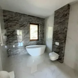 Nowoczesna łazienka z marmurowymi płytkami na ścianach, wolnostojącą białą wanną i podwieszaną toaletą, okno z widokiem na zieleń.