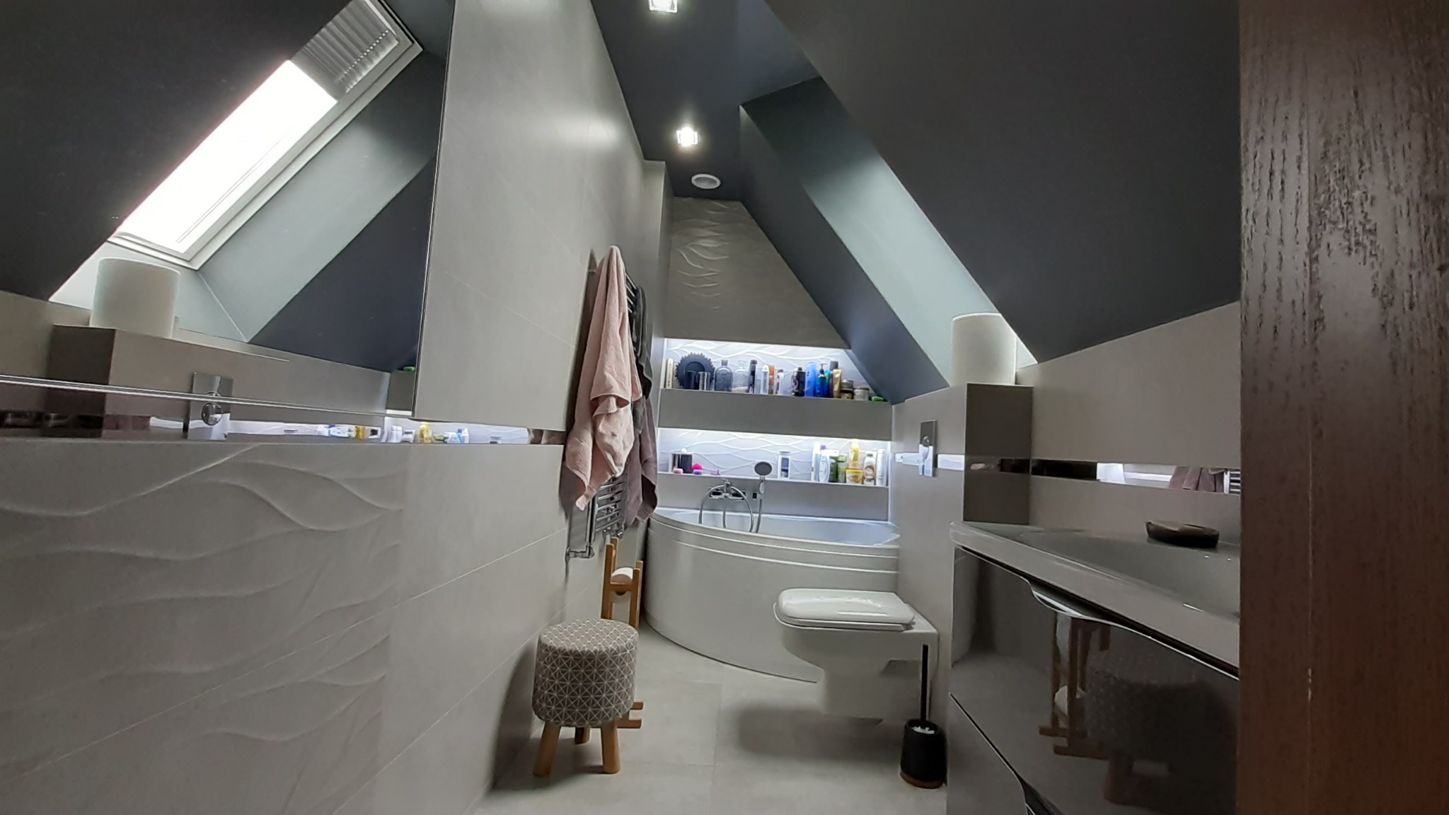 Nowoczesna łazienka na poddaszu z oknem dachowym, szarą kolorystyką, wanną narożną, toaletą i dekoracyjną ścianą z falistą strukturą.