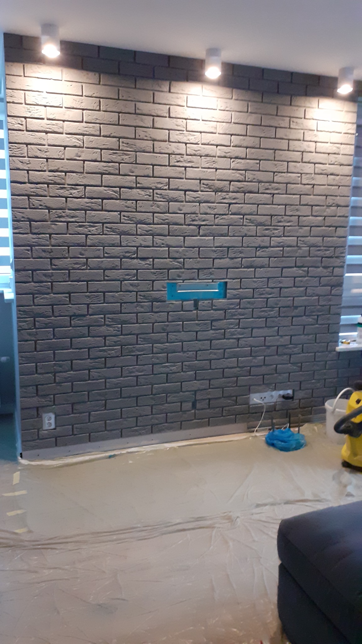 Ściana wyłożona szarą cegłą dekoracyjną z widocznym otworem na instalację, zabezpieczona folią podłoga podczas prac wykończeniowych.