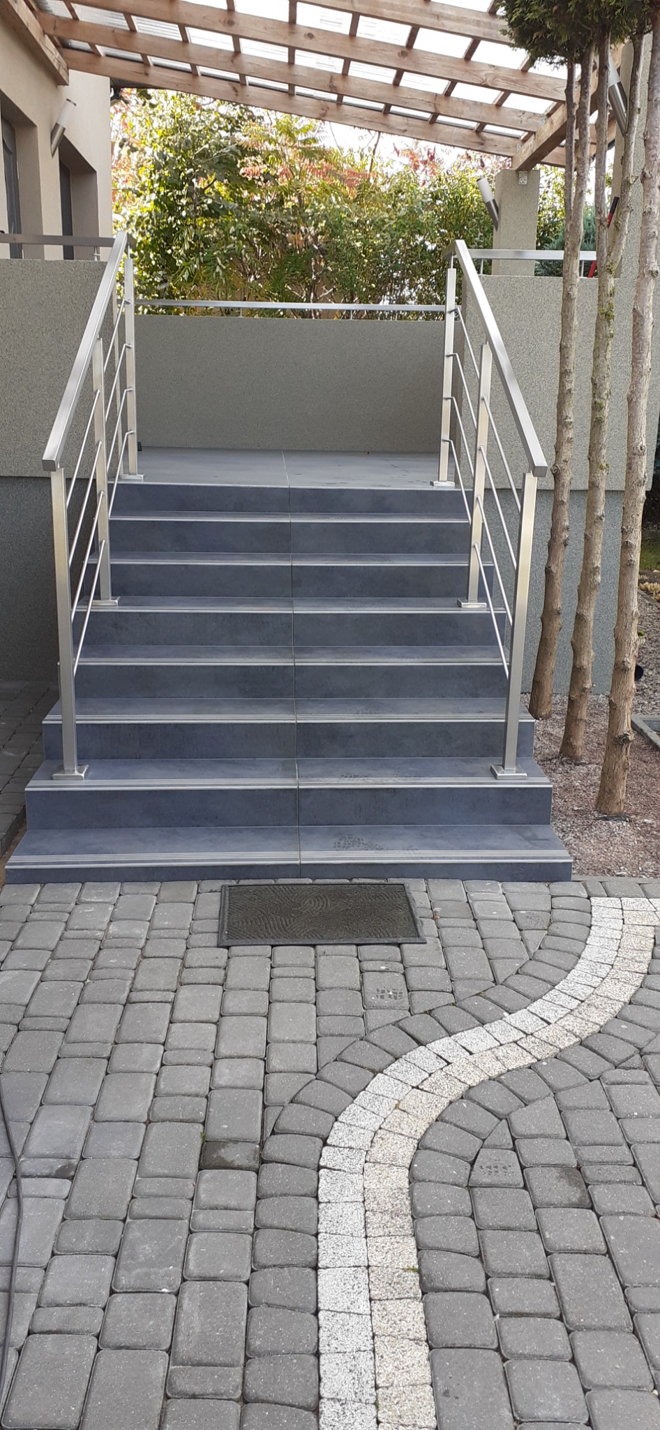 Zewnętrzne schody wyłożone szarymi płytkami z metalową balustradą, prowadzące do budynku z szarą elewacją, na dole kostka brukowa z jasnym łukiem.