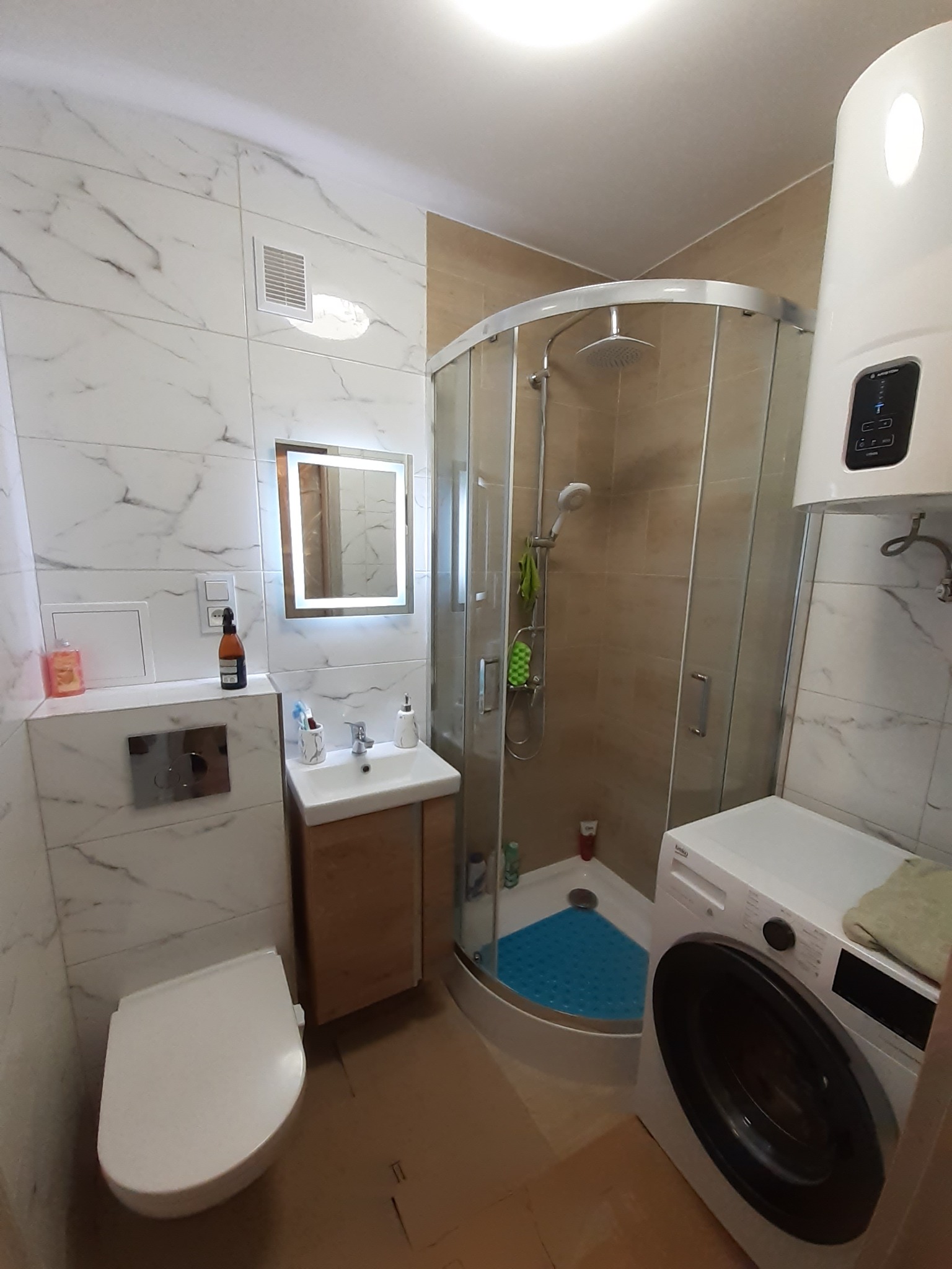 Ukończona aranżacja łazienki z kabiną prysznicową, umywalką z szafką, toaletą i pralką, wykończona białymi płytkami imitującymi marmur i beżowymi płytkami w kabinie prysznicowej.