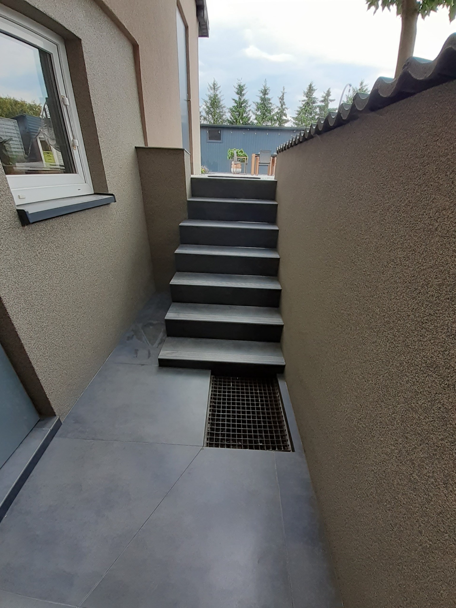 Zewnętrzne schody wyłożone szarymi płytkami prowadzące w dół, ściany z beżowym tynkiem strukturalnym, widoczna kratka ściekowa i okno z białymi ramami.