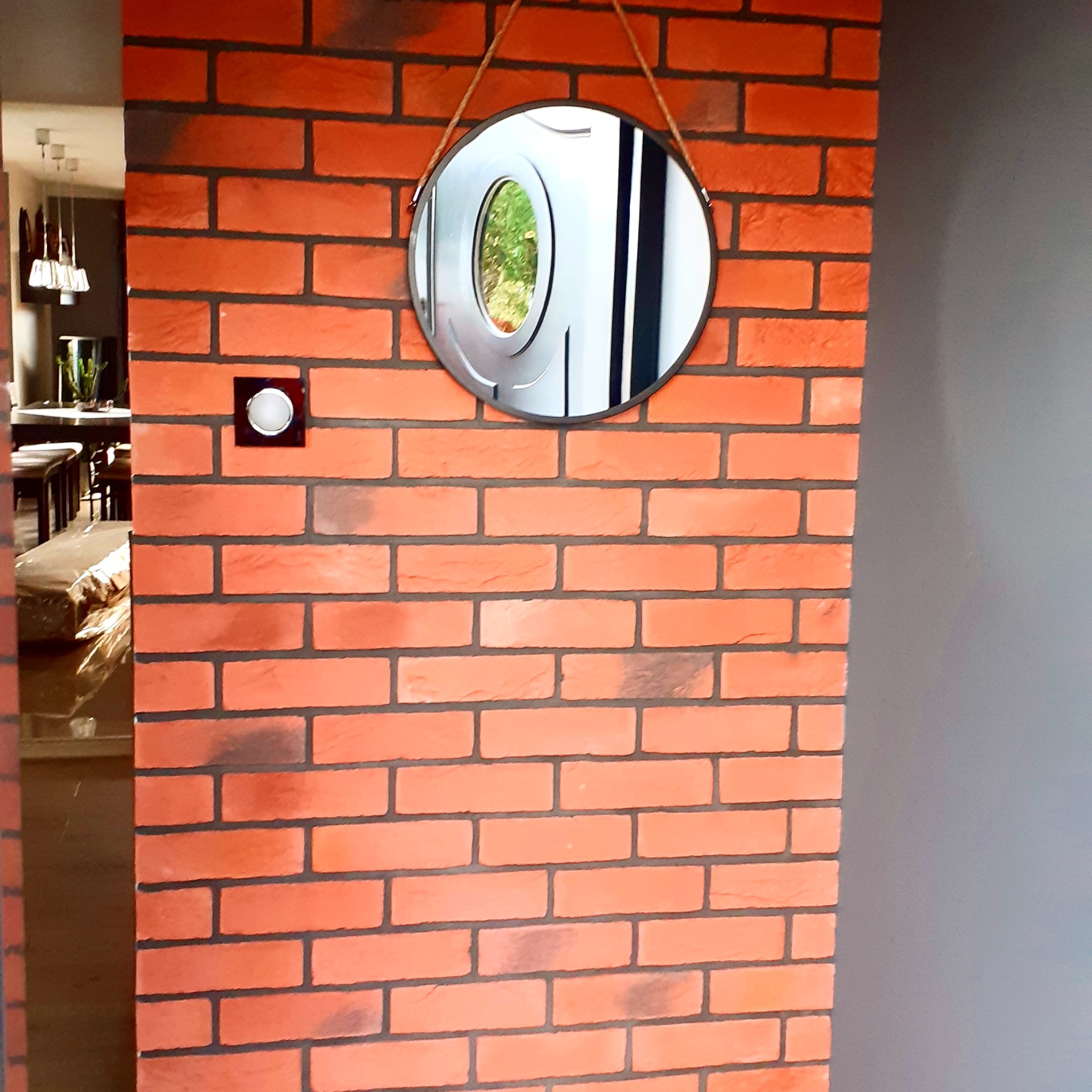 Ściana wykończona czerwoną cegłą klinkierową z widocznym włącznikiem światła i okrągłym lustrem wiszącym na sznurku, fragment widoku na wnętrze pomieszczenia.
