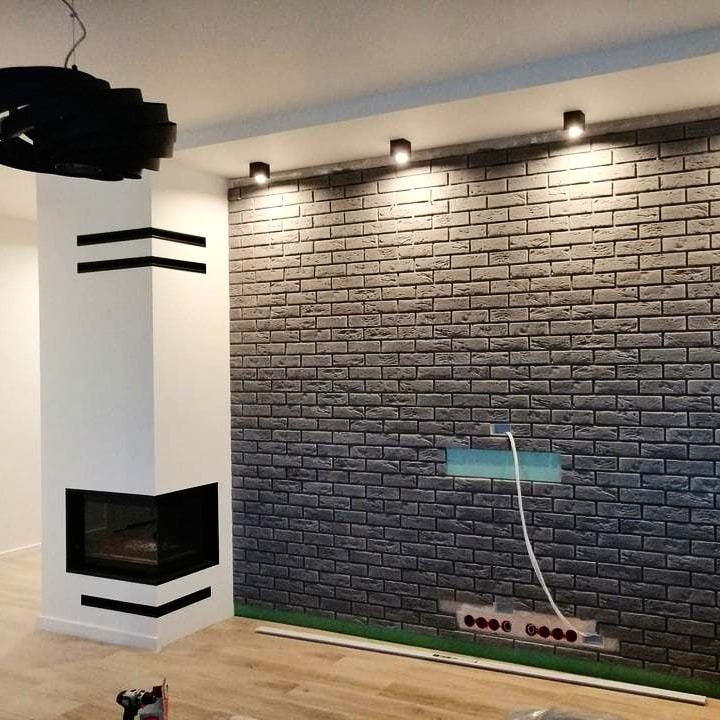 Szara ściana z cegły dekoracyjnej z oświetleniem punktowym, widoczne gniazdka elektryczne i biały kominek narożny z czarnymi akcentami w nowoczesnym wnętrzu.