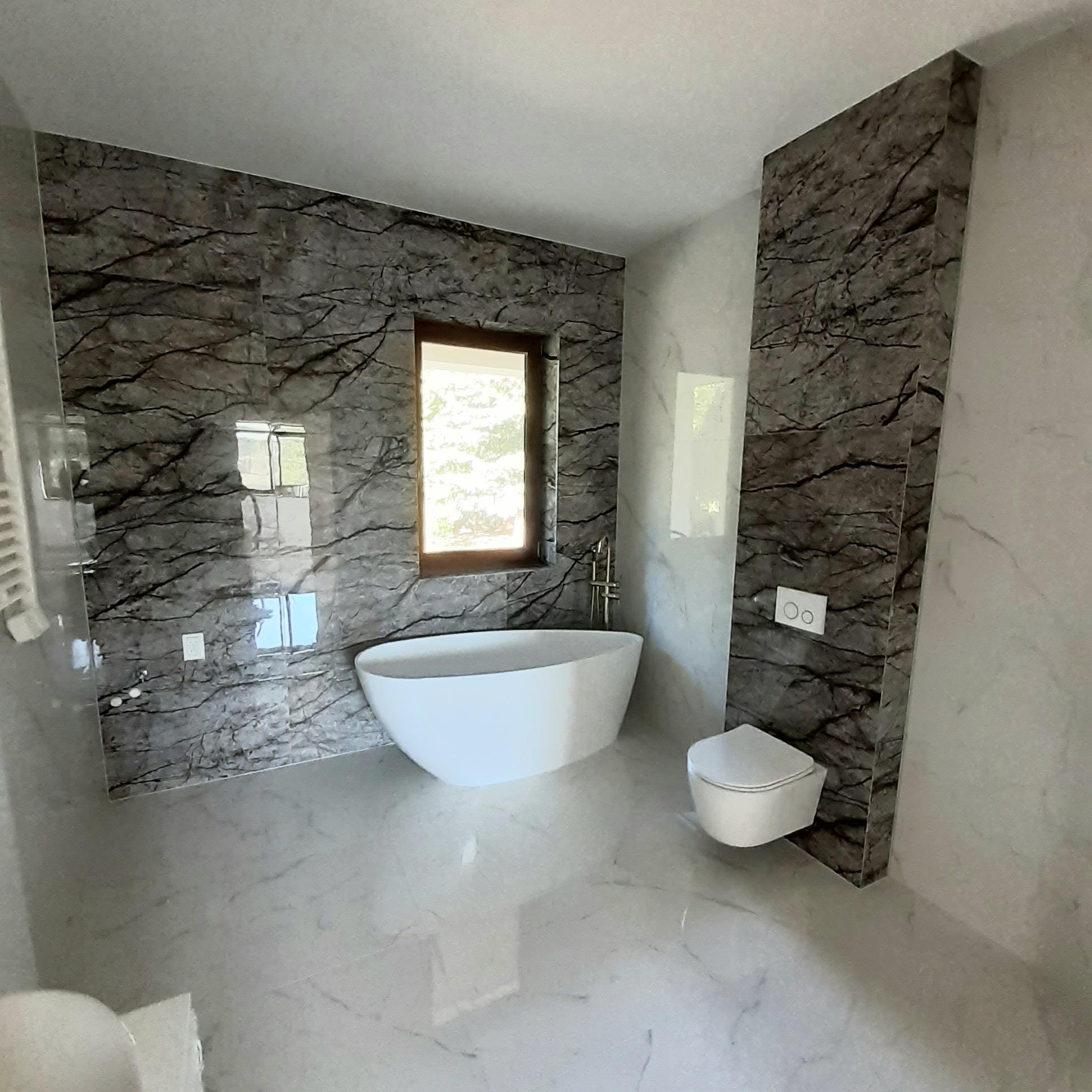 Nowoczesna łazienka z marmurowymi płytkami na ścianach, wolnostojącą białą wanną i podwieszaną toaletą, okno z widokiem na zieleń.