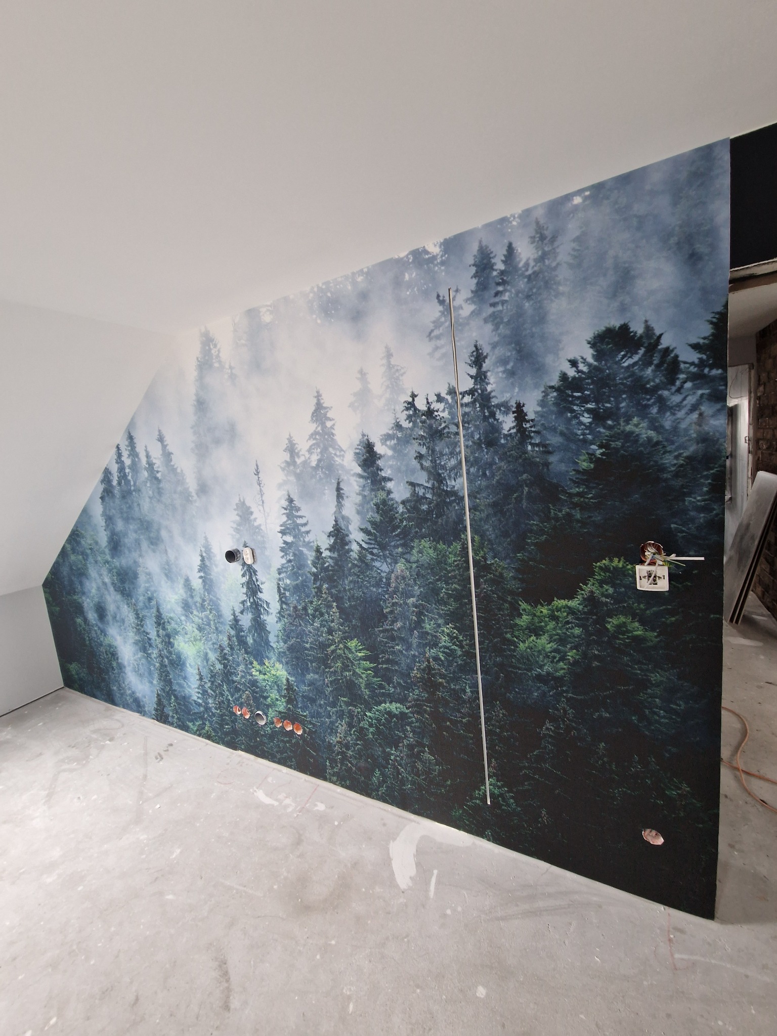 Fototapeta z motywem mglistego lasu iglastego na ścianie ze skosem, widoczne gniazdka elektryczne i wystające kable.