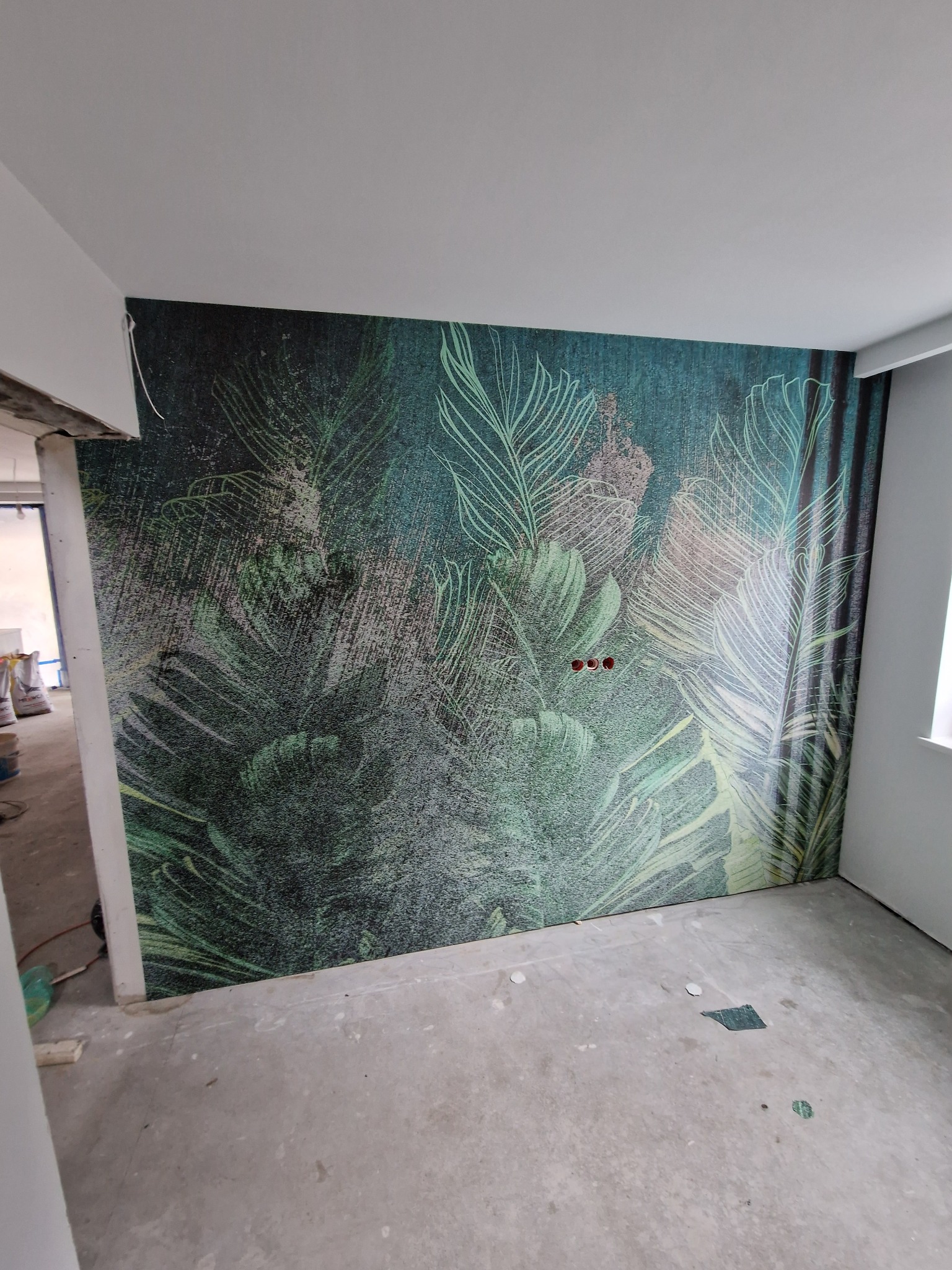 Ściana pokryta tapetą z motywem tropikalnych liści w odcieniach zieleni i szarości; widoczne gniazdka elektryczne i surowy beton na podłodze.