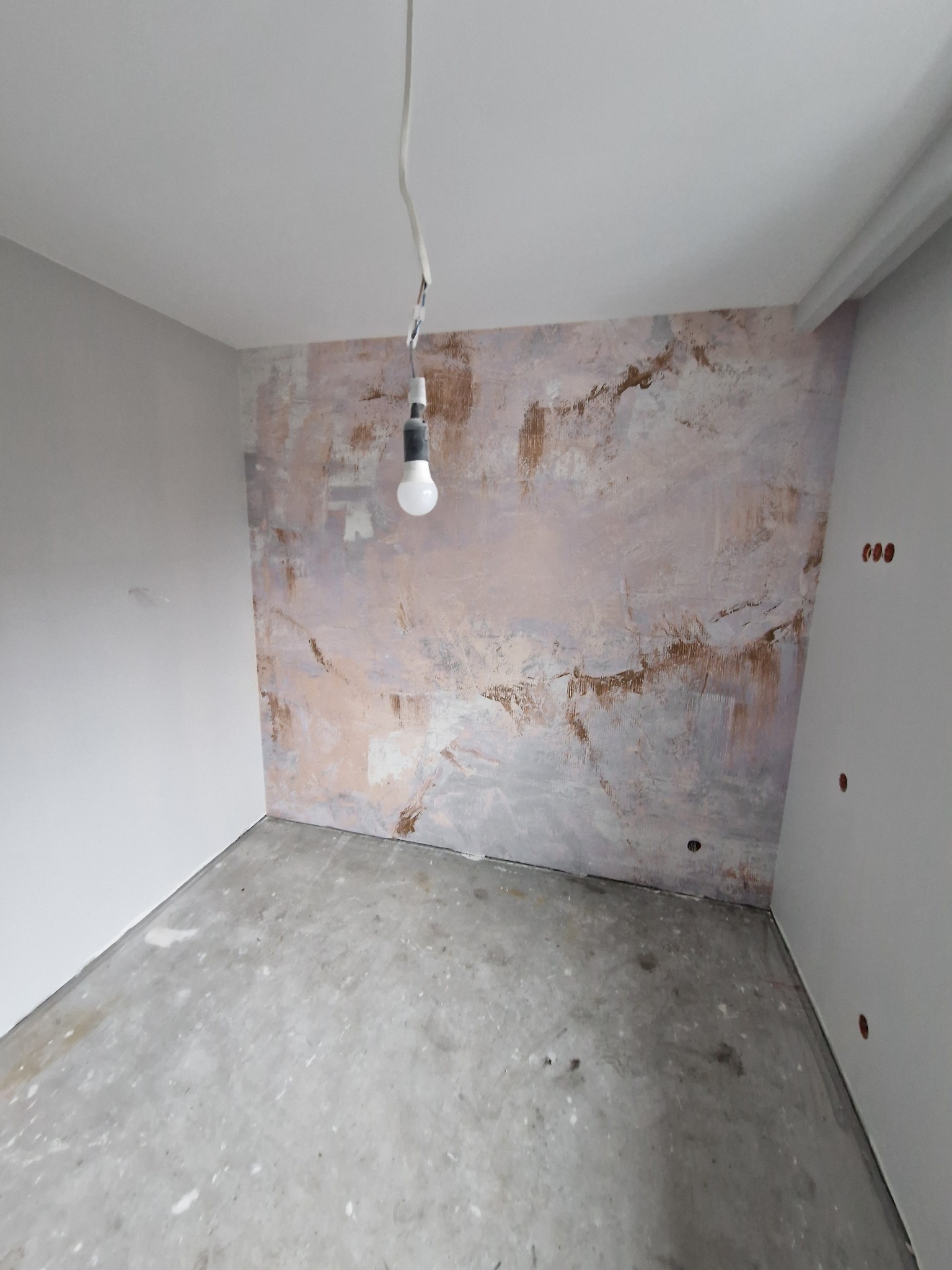 Przygotowanie ściany do prac wykończeniowych: zaszpachlowana ściana z widocznymi śladami po poprzednich warstwach, surowy beton na podłodze, wisząca żarówka na kablu.