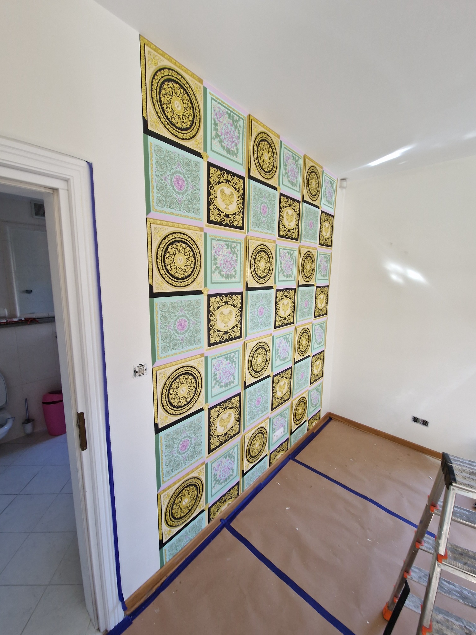 Ściana w pokoju z wzorzystą tapetą w kwadraty, zabezpieczona folią i taśmą malarską podczas prac remontowych, widoczna drabina.