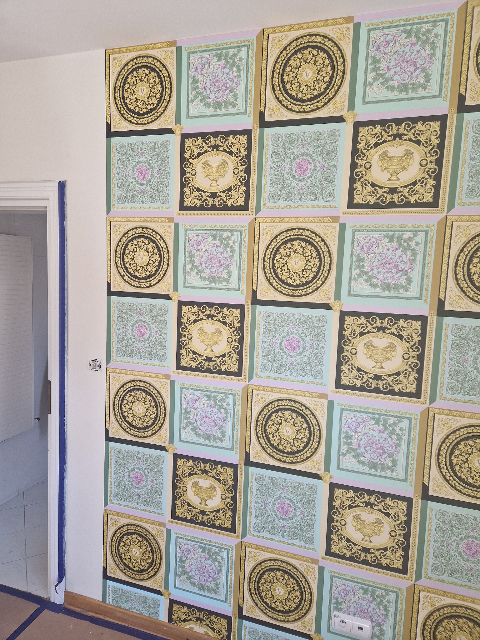 Fragment ściany pokrytej luksusową tapetą w kwadratowe wzory z motywami Versace, widoczne niedokończone prace wykończeniowe przy drzwiach.