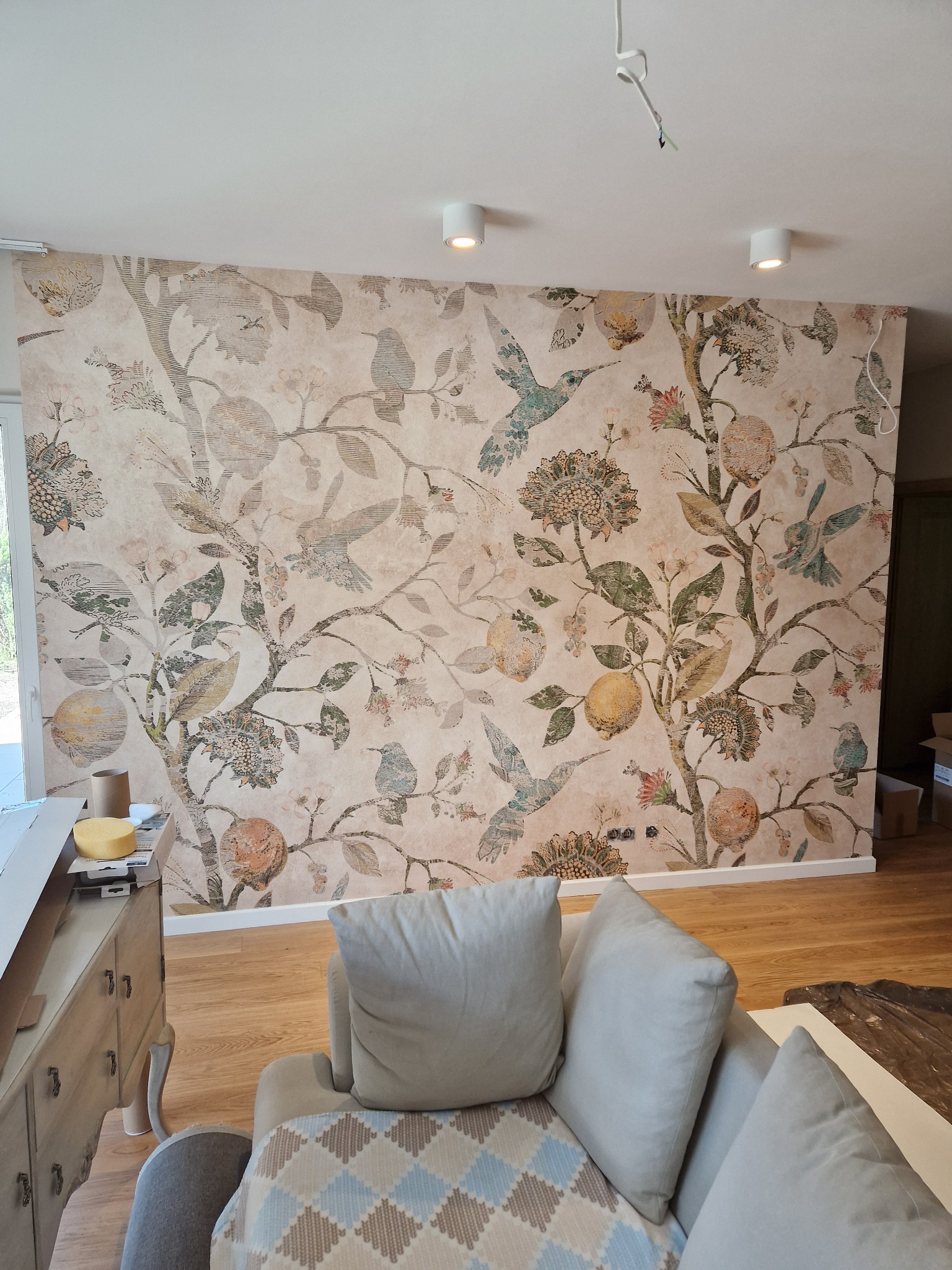 Ściana pokryta beżową tapetą w motywy roślinne i ptasie, widoczne kolibry i owoce cytrusowe, oświetlenie sufitowe, fragment sofy w dolnej części kadru.