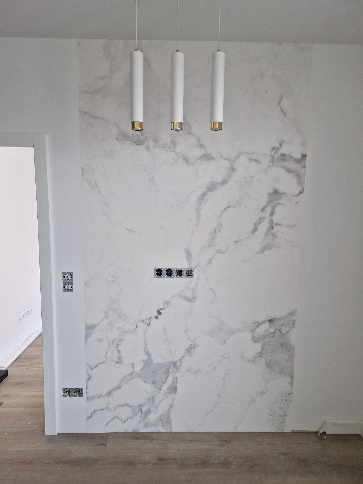 Ściana w Warszawie z tapetą imitującą marmur, podświetlona trzema cylindrycznymi lampami wiszącymi, gniazdka elektryczne zamontowane w ścianie.