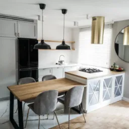 Nowoczesna kuchnia z drewnianym stołem i szarymi krzesłami, oświetlona czarnymi lampami wiszącymi, z widoczną zabudową kuchenną w odcieniach szarości i złotym okapem cylindrycznym.