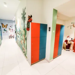Kolorowy korytarz z szafkami w odcieniach czerwieni i niebieskiego, dekorowany malowidłami ściennymi z motywami zwierząt i roślin tropikalnych, pluszowy miś siedzący na szafce, oraz dekoracja...