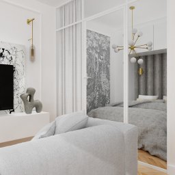 Home by Koja - Nowoczesna aranżacja salonu z widokiem na sypialnię przez szklaną ścianę, szary narożnik na pierwszym planie, parkiet w jodełkę, abstrakcyjna grafika na ścianie, złota lampa ścienna.