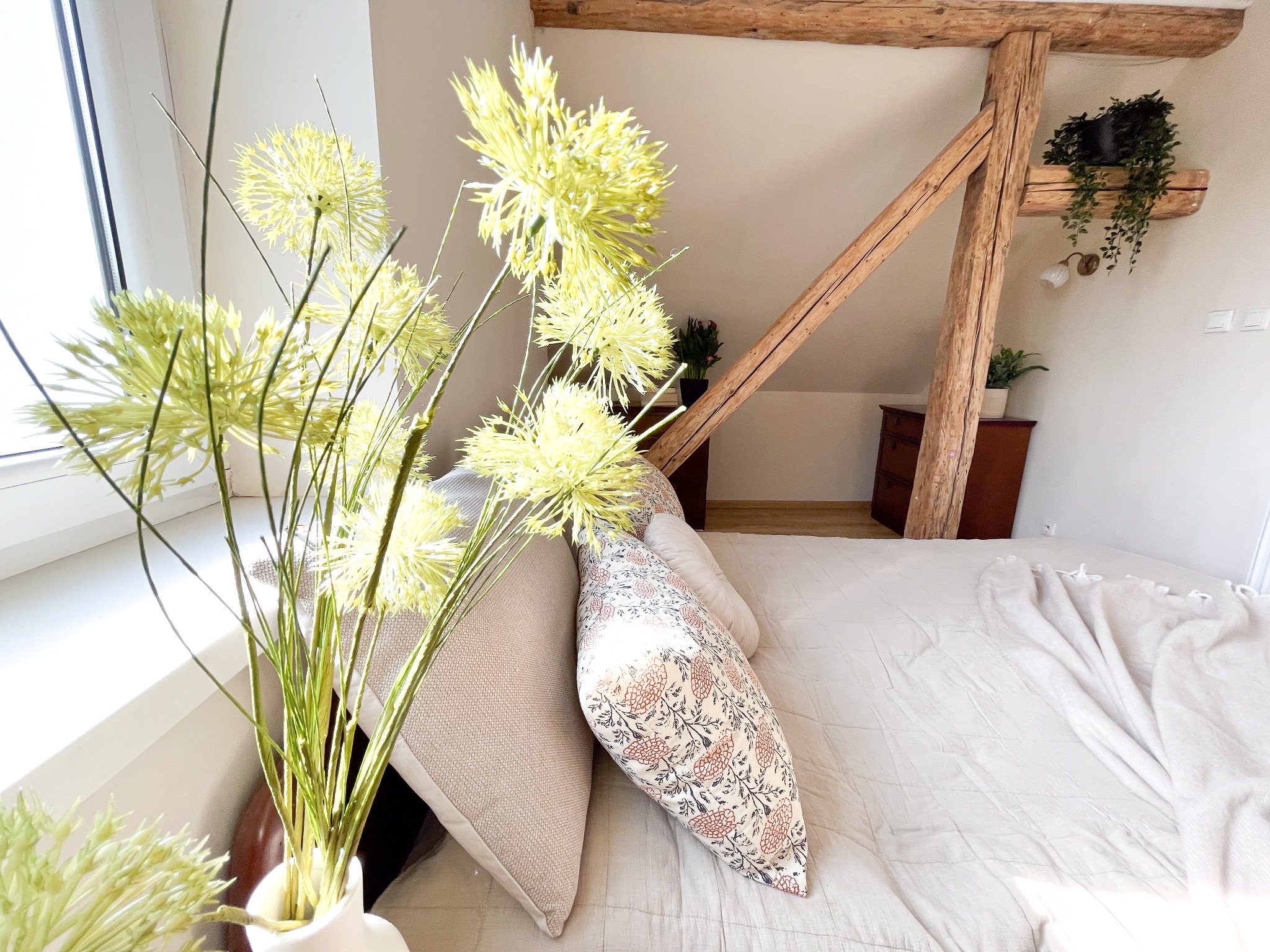 Jasny pokój na poddaszu z drewnianą konstrukcją stropu, łóżkiem z beżową pościelą i dekoracyjną rośliną w wazonie na parapecie.