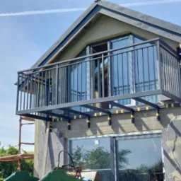 Montaż stalowej balustrady balkonowej w nowo budowanym domu z widocznymi śladami prac budowlanych i rusztowaniem w tle.