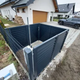 Szara, lamelowa obudowa śmietnika z metalu, częściowo zmontowana, widoczna na tle nowoczesnego domu jednorodzinnego z drewnem na opał i szarym SUV-em na podjeździe. Wokół widoczne ślady prac...