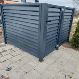 Nowoczesna, grafitowa obudowa śmietnika z poziomych listew, zamontowana na kostce brukowej przed domem. Widoczny zamek z kluczem oraz tarcza do szlifowania leżąca na ziemi.