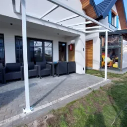 Zadaszenie tarasu z białej konstrukcji stalowej, z widocznymi elementami mocującymi do podłoża i śrubami, na tle domu z niebieskimi okiennicami i kostką brukową na tarasie.