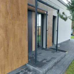 Nowoczesne wejście do domu z przeszklonym zadaszeniem na metalowej konstrukcji, elewacja z drewnopodobnym wykończeniem, szara kostka brukowa przed drzwiami.