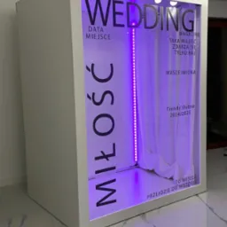 Biała fotobudka z fioletowym oświetleniem i napisem 'Miłość' w Rakowiskach, prezentująca motyw ślubny z elementami dekoracyjnymi i informacjami o dacie i miejscu.