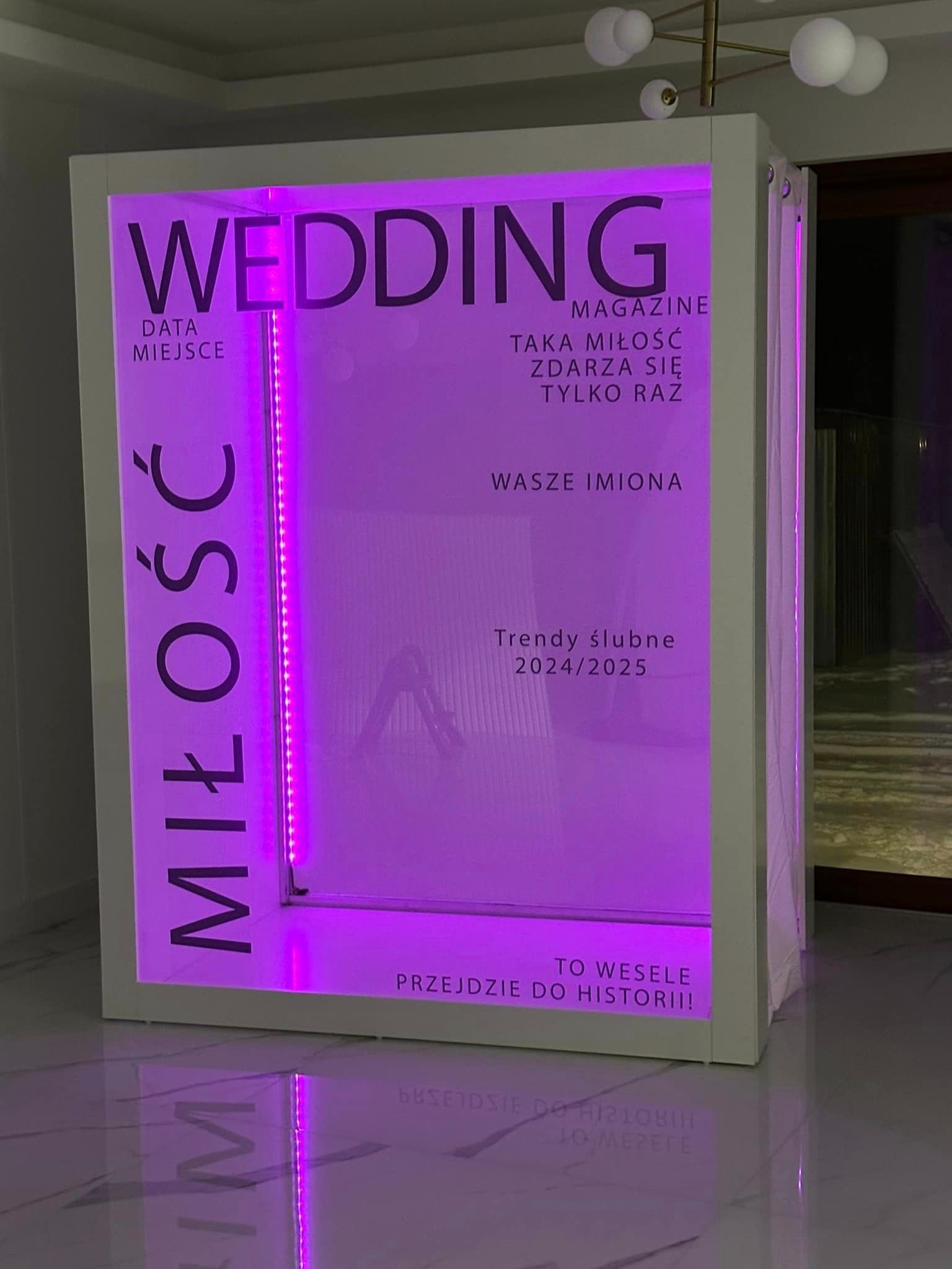 Biała fotobudka w kształcie okładki magazynu ślubnego z fioletowym podświetleniem LED, napisy: WEDDING, MIŁOŚĆ, Trendy ślubne 2024/2025.