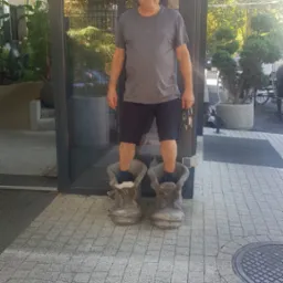 Mężczyzna stoi wewnątrz dwóch dużych, betonowych donic w kształcie butów, trzymając pęk kluczy w dłoni, na tle przeszklonego wejścia do budynku.