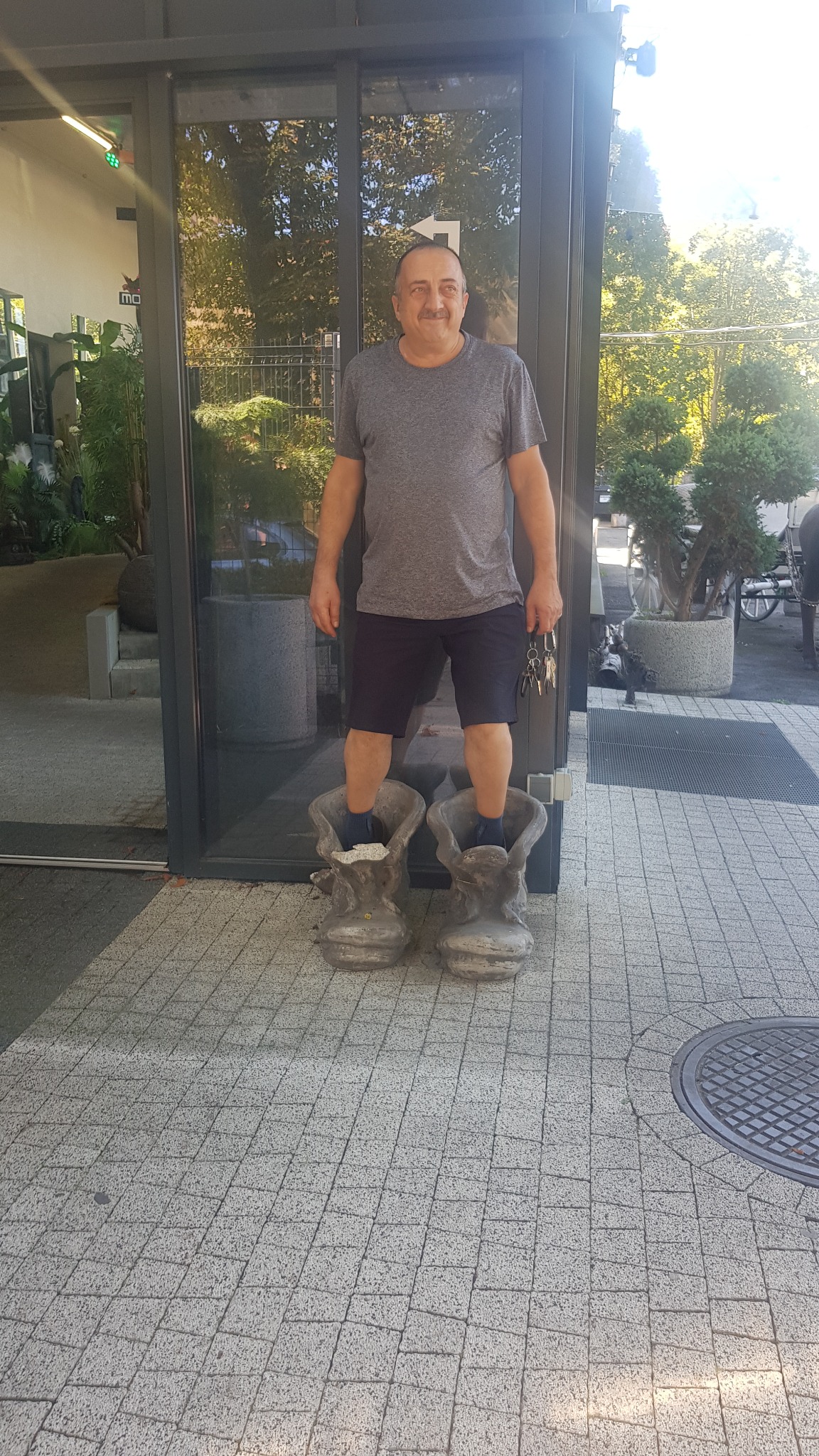 Mężczyzna stoi wewnątrz dwóch dużych, betonowych donic w kształcie butów, trzymając pęk kluczy w dłoni, na tle przeszklonego wejścia do budynku.