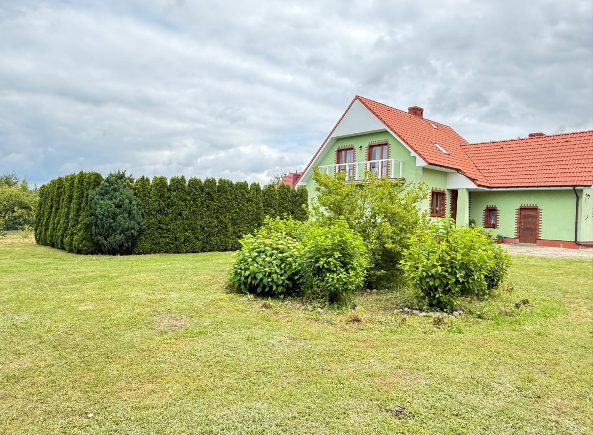 Zielony dom z czerwonym dachem i balkonem, otoczony bujną zielenią i wysokim żywopłotem, na tle pochmurnego nieba. Ujęcie z poziomu trawnika.
