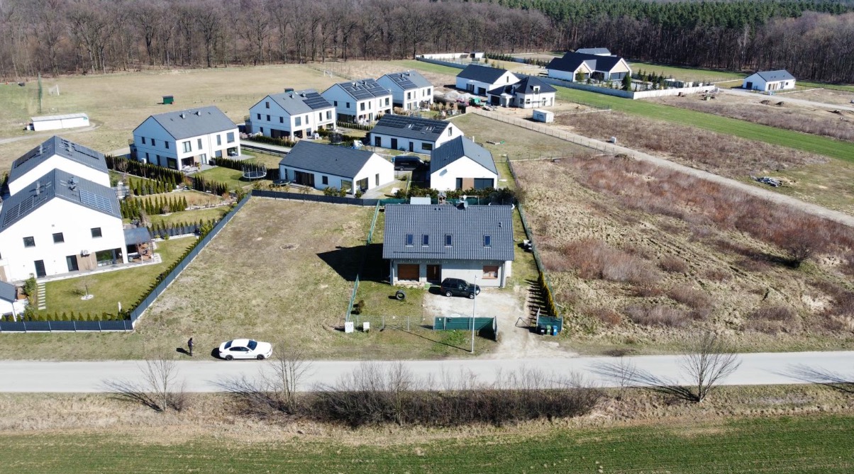 Zdjęcie z drona osiedla domów jednorodzinnych. Na pierwszym planie dom z szarym dachem i podjazdem, w tle widoczne inne domy i las.