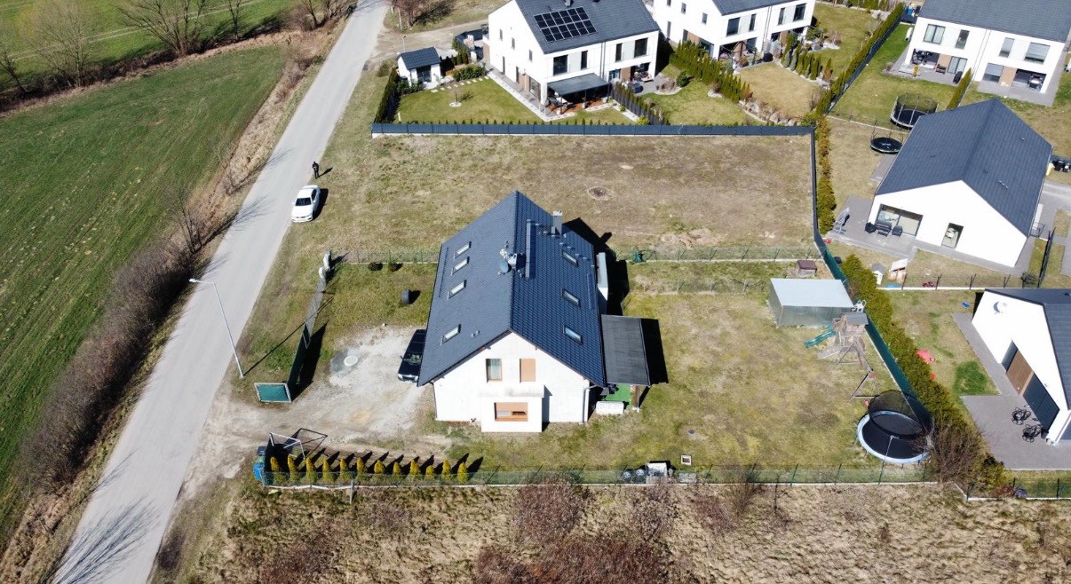 Zdjęcie z drona: nowoczesny dom z ciemnym dachem, białą elewacją i dużym ogrodem w otoczeniu innych domów jednorodzinnych.