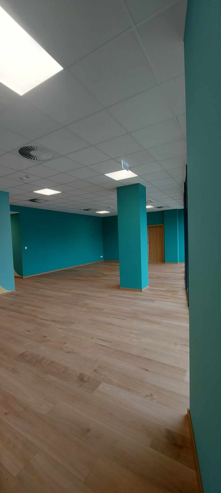 Wnętrze biura po remoncie z turkusowymi ścianami, jasną podłogą z paneli i białym sufitem podwieszanym z kwadratowymi lampami.