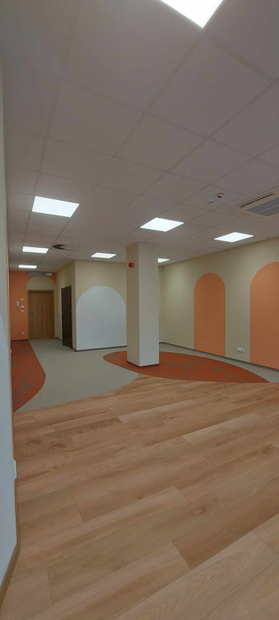 Jasne, przestronne wnętrze biurowe z drewnianą podłogą, pomalowanymi na pomarańczowo wnękami w ścianach i podwieszanym sufitem z kwadratowymi panelami oświetleniowymi.