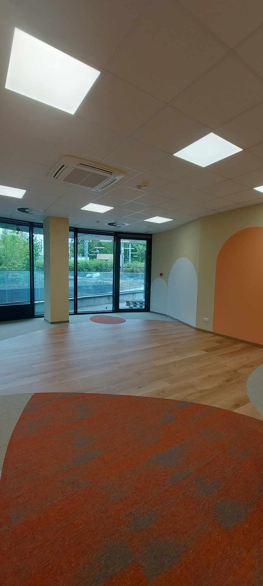 Jasne, przestronne wnętrze biurowe z drewnianą podłogą i pomarańczowymi akcentami na ścianach, widok na zewnątrz przez duże okna.