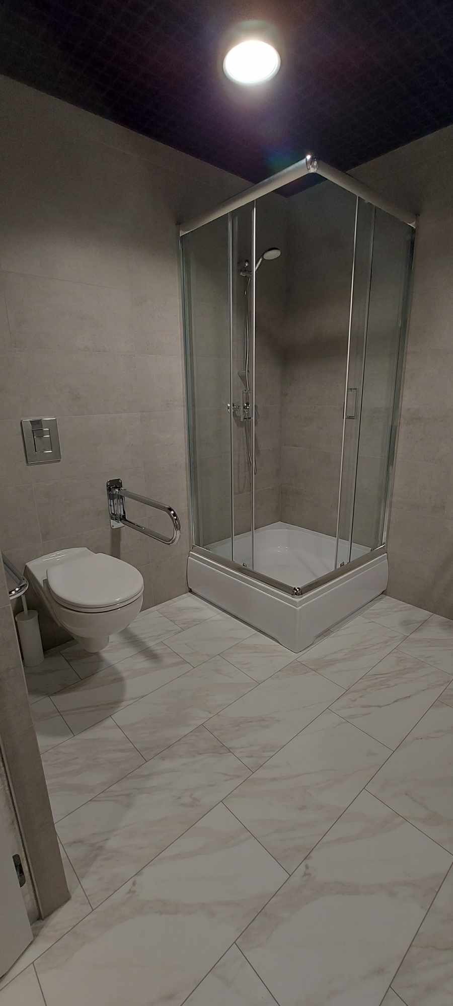 Wykończona łazienka z kabiną prysznicową narożną, wiszącą toaletą z uchwytem dla niepełnosprawnych, płytkami podłogowymi imitującymi marmur i szarą glazurą ścienną.
