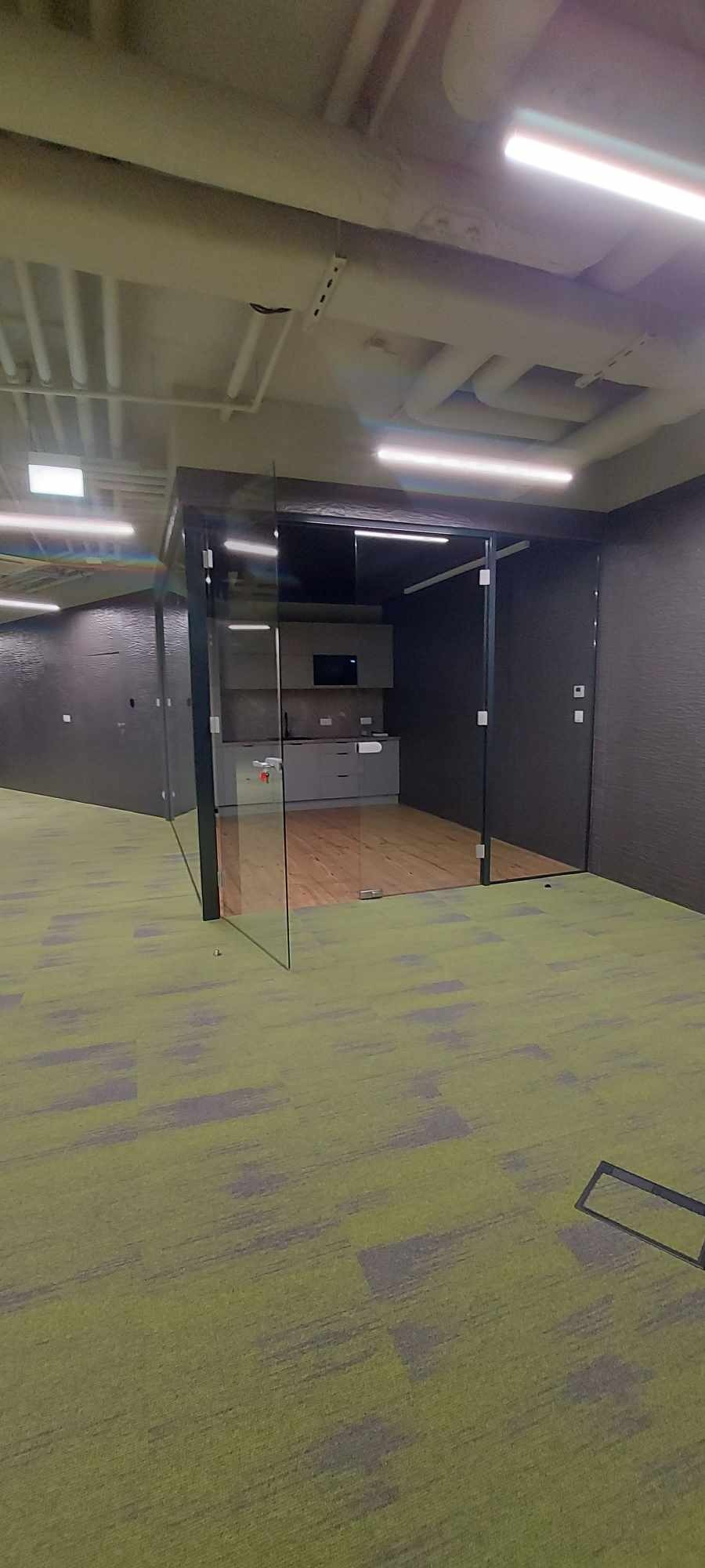 Wnętrze biura z przeszkloną ścianą działową i widoczną kuchnią w tle, zielona wykładzina podłogowa.