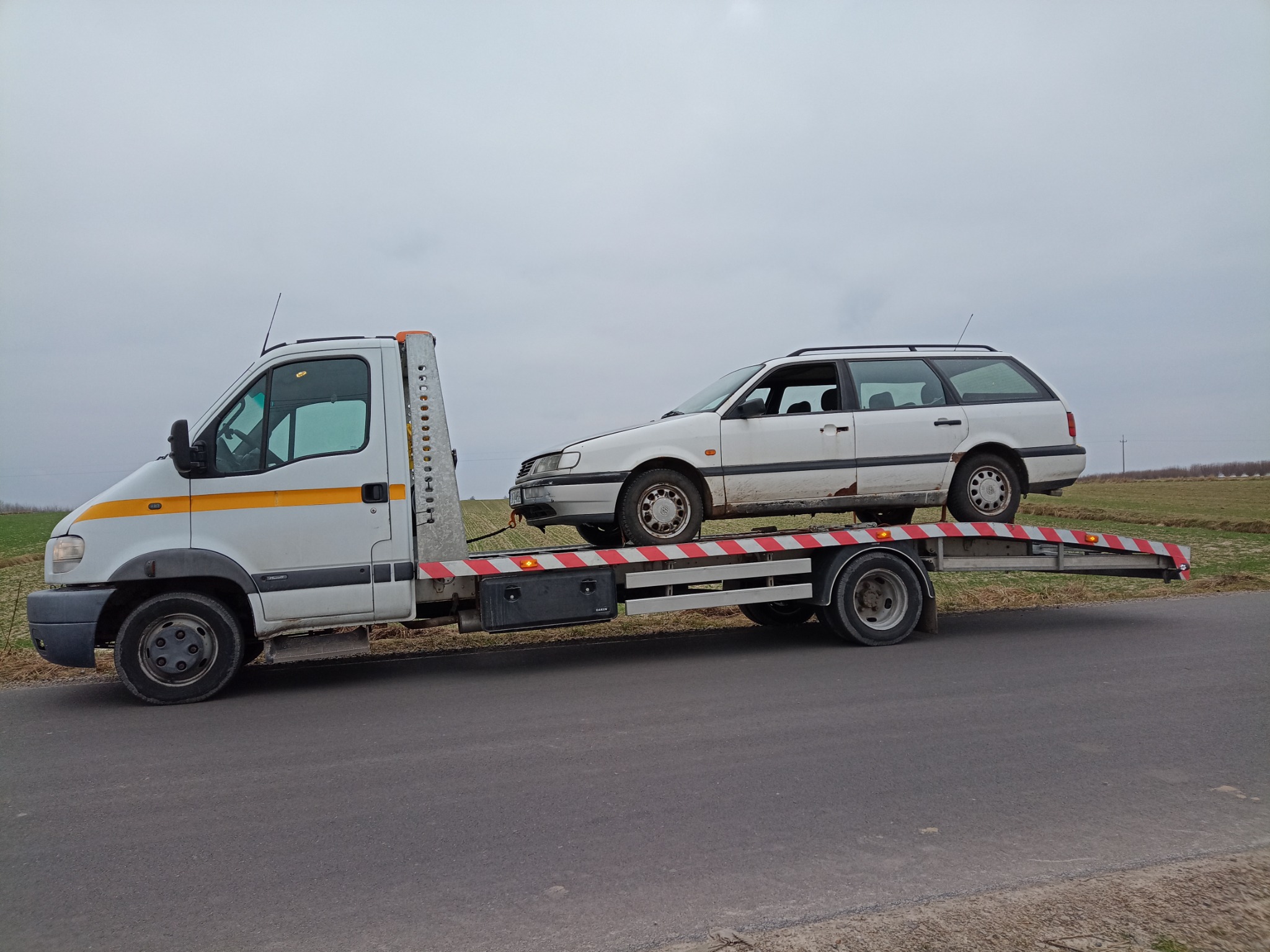 Biała platforma transportowa z załadowanym białym samochodem kombi na tle szarego nieba i pola.