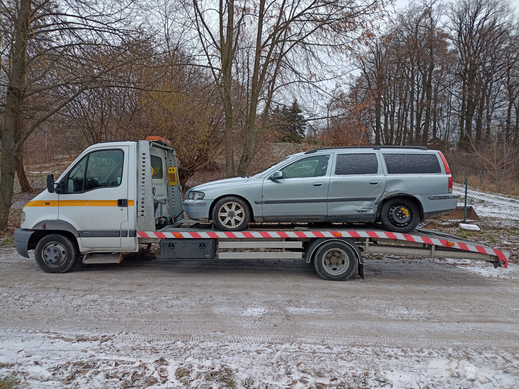 Uszkodzony srebrny samochód kombi przewożony na białej ciężarówce z żółtym pasem, na tle zimowego krajobrazu z drzewami bez liści.