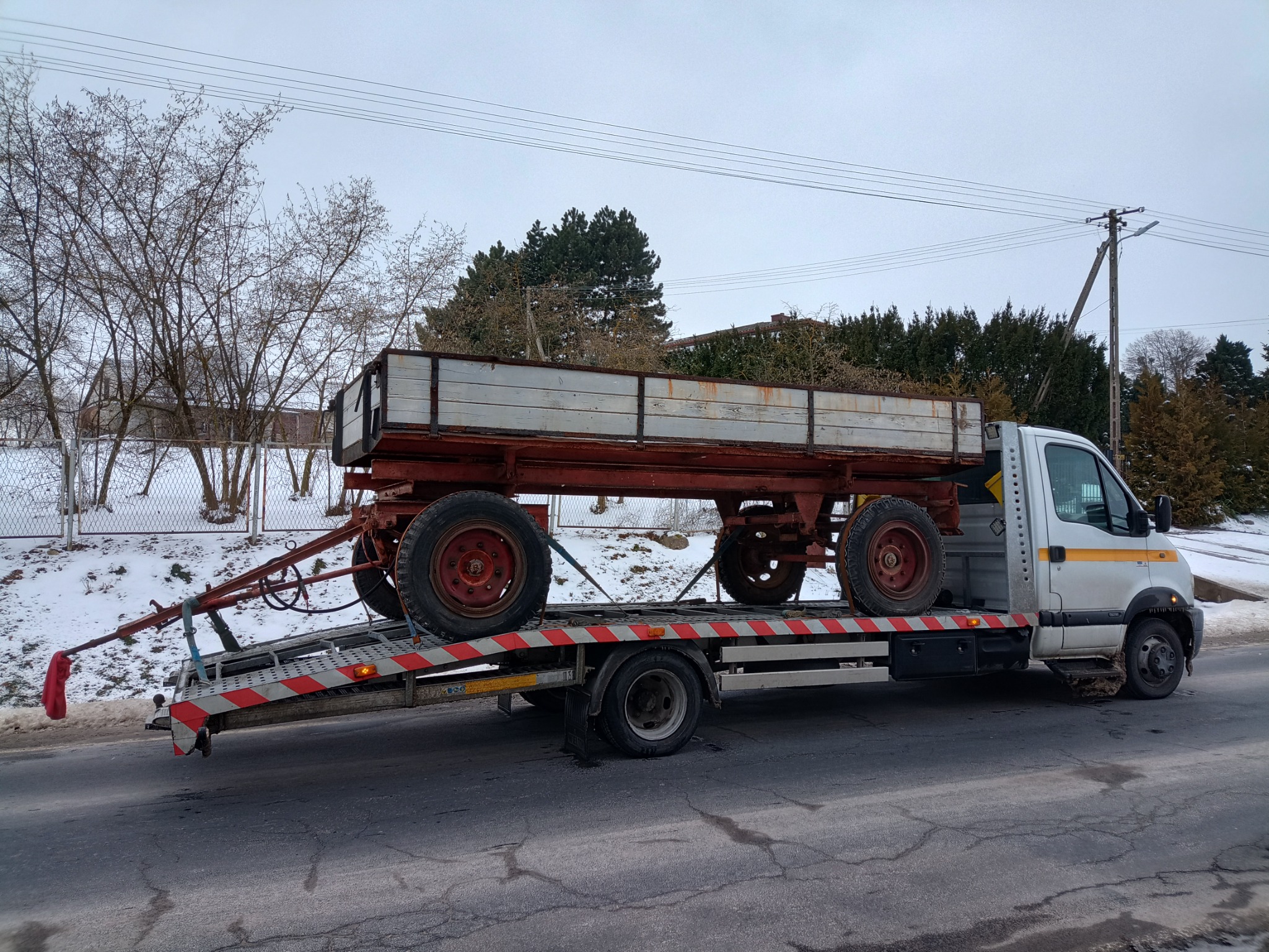 Przyczepa rolnicza z drewnianymi burtami transportowana na platformie pojazdu pomocy drogowej w zimowej scenerii.