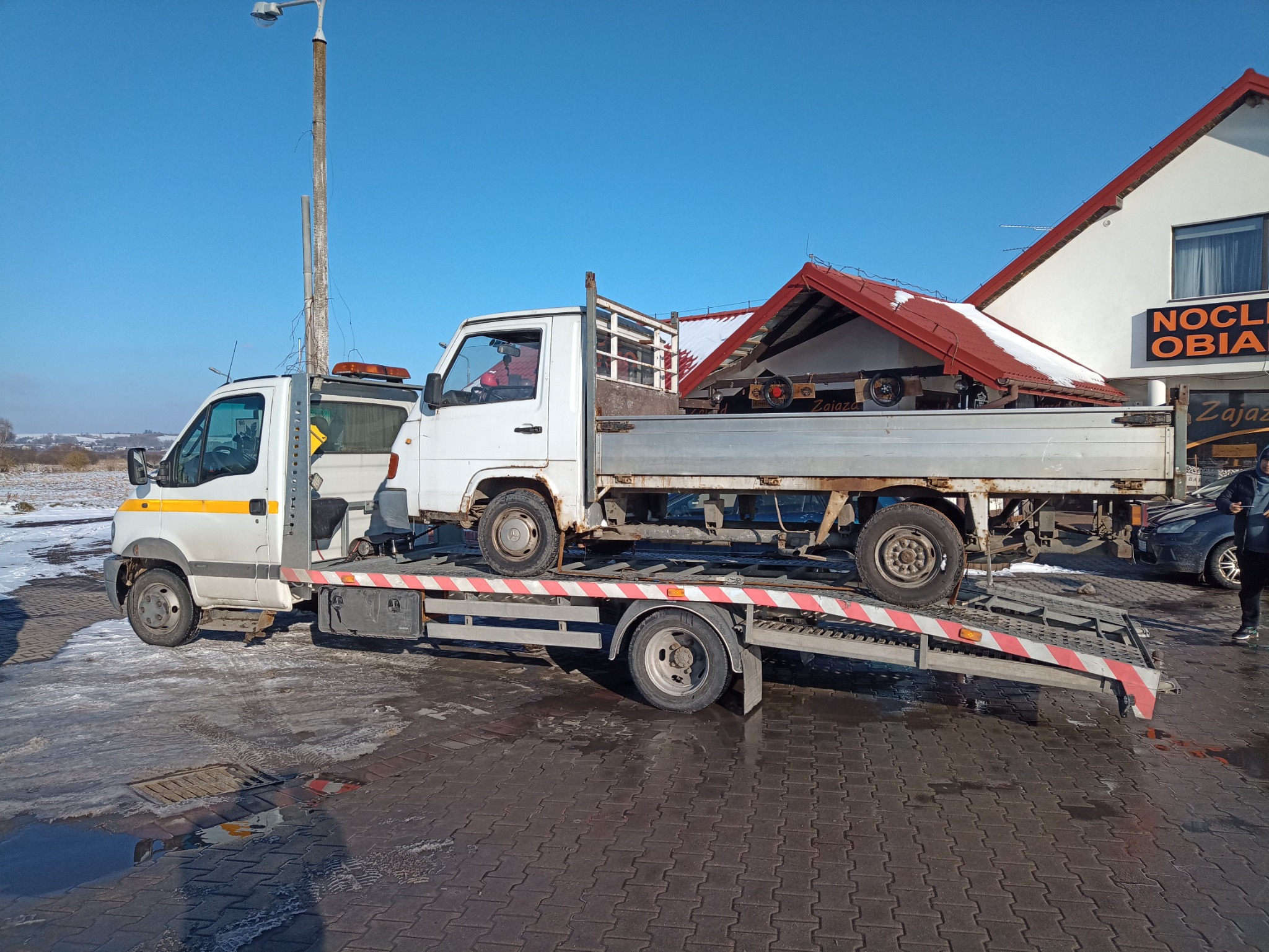 Biała laweta z żółtymi pasami transportuje uszkodzony, biały samochód dostawczy na tle zimowego krajobrazu i budynku z napisem 'NOCLEGI OBIADY'.