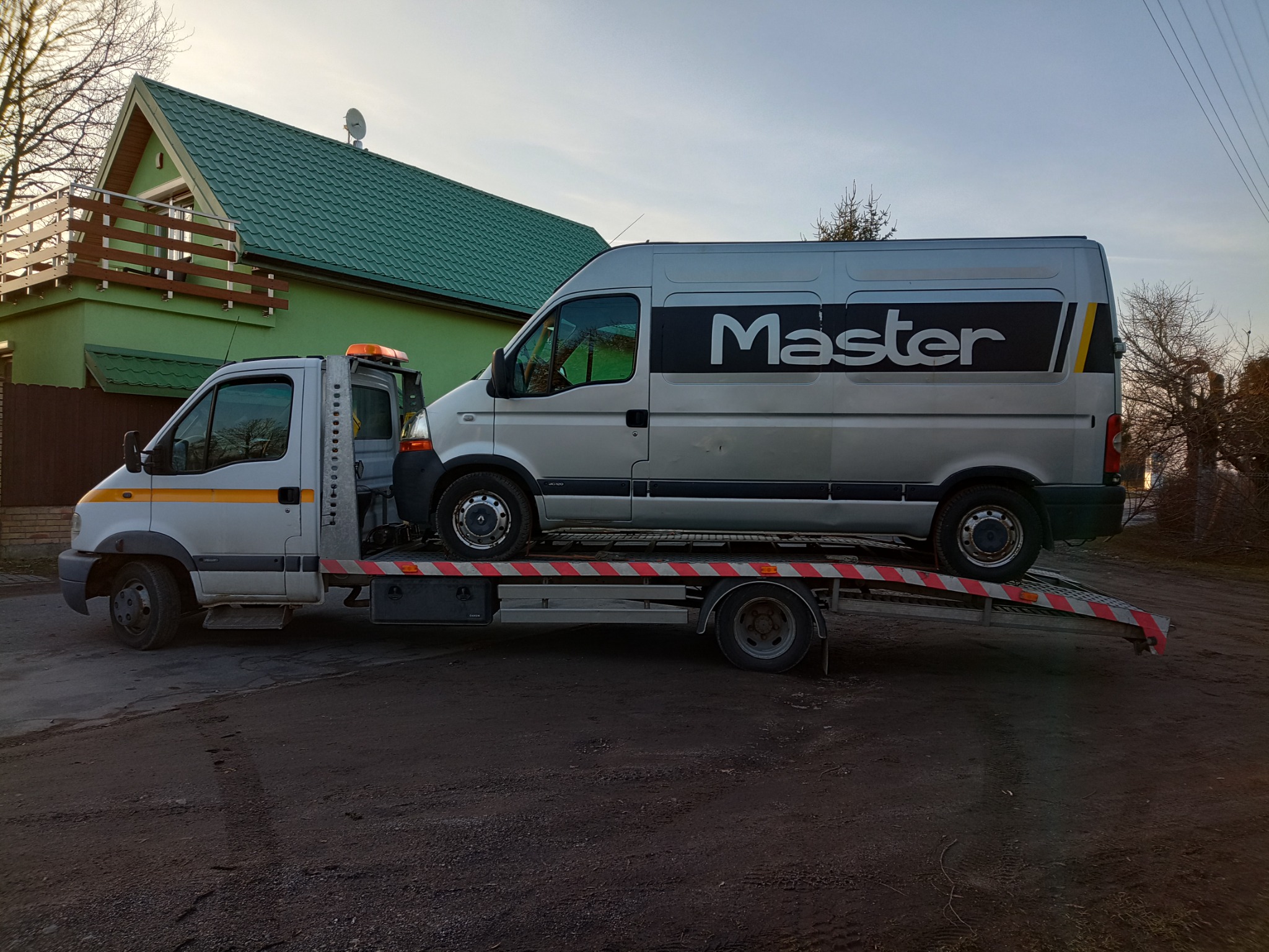 Srebrny bus marki Master przewożony na pojeździe pomocy drogowej, z zielonym domem w tle.