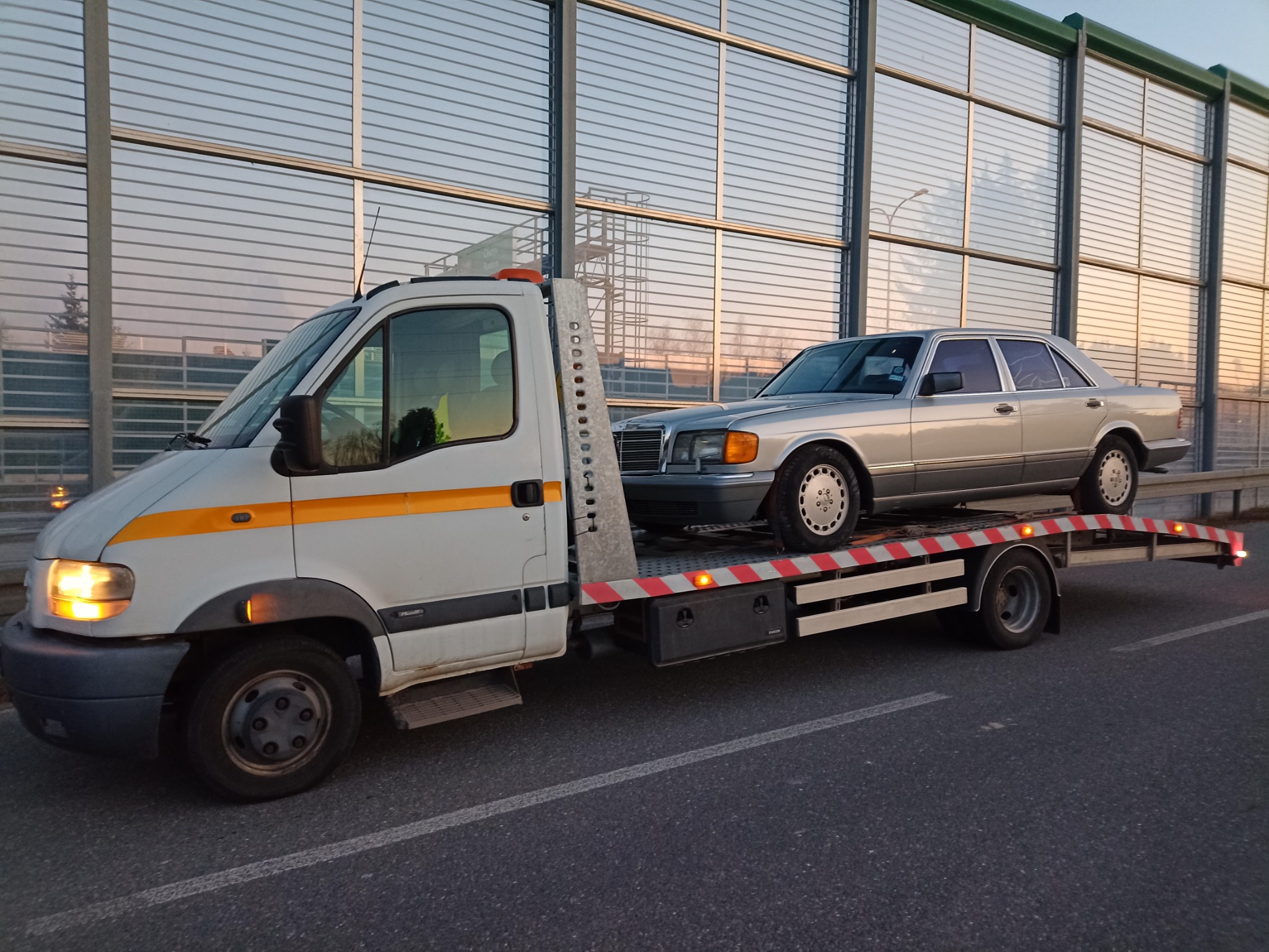 Srebrny Mercedes W126 transportowany na białej platformie pojazdu pomocy drogowej z pomarańczowymi pasami, na tle ekranów akustycznych przy autostradzie.