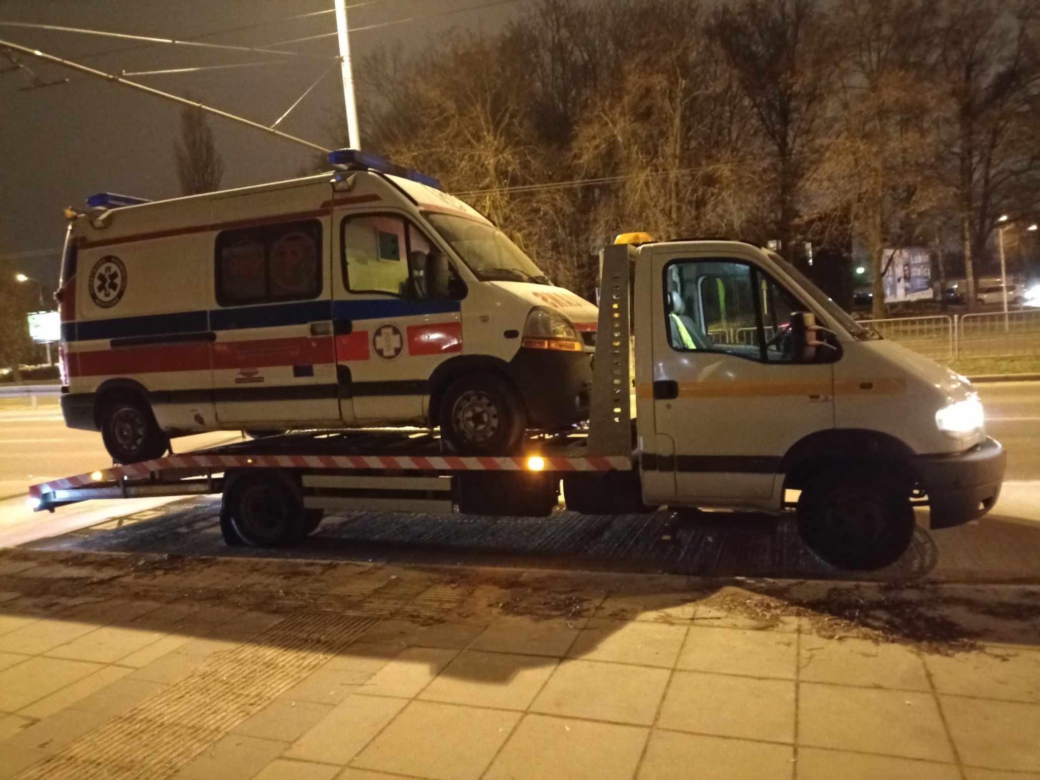 Ambulans transportowany na platformie pomocy drogowej w warunkach nocnych, widoczne światła ostrzegawcze i elementy otoczenia miejskiego.