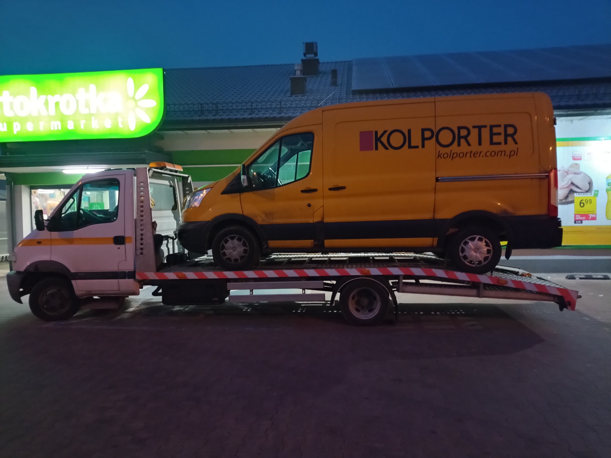 Biało-żółta platforma transportowa z żółtym vanem 'KOLPORTER' na pokładzie, zaparkowana przed sklepem 'Stokrotka' wieczorem.