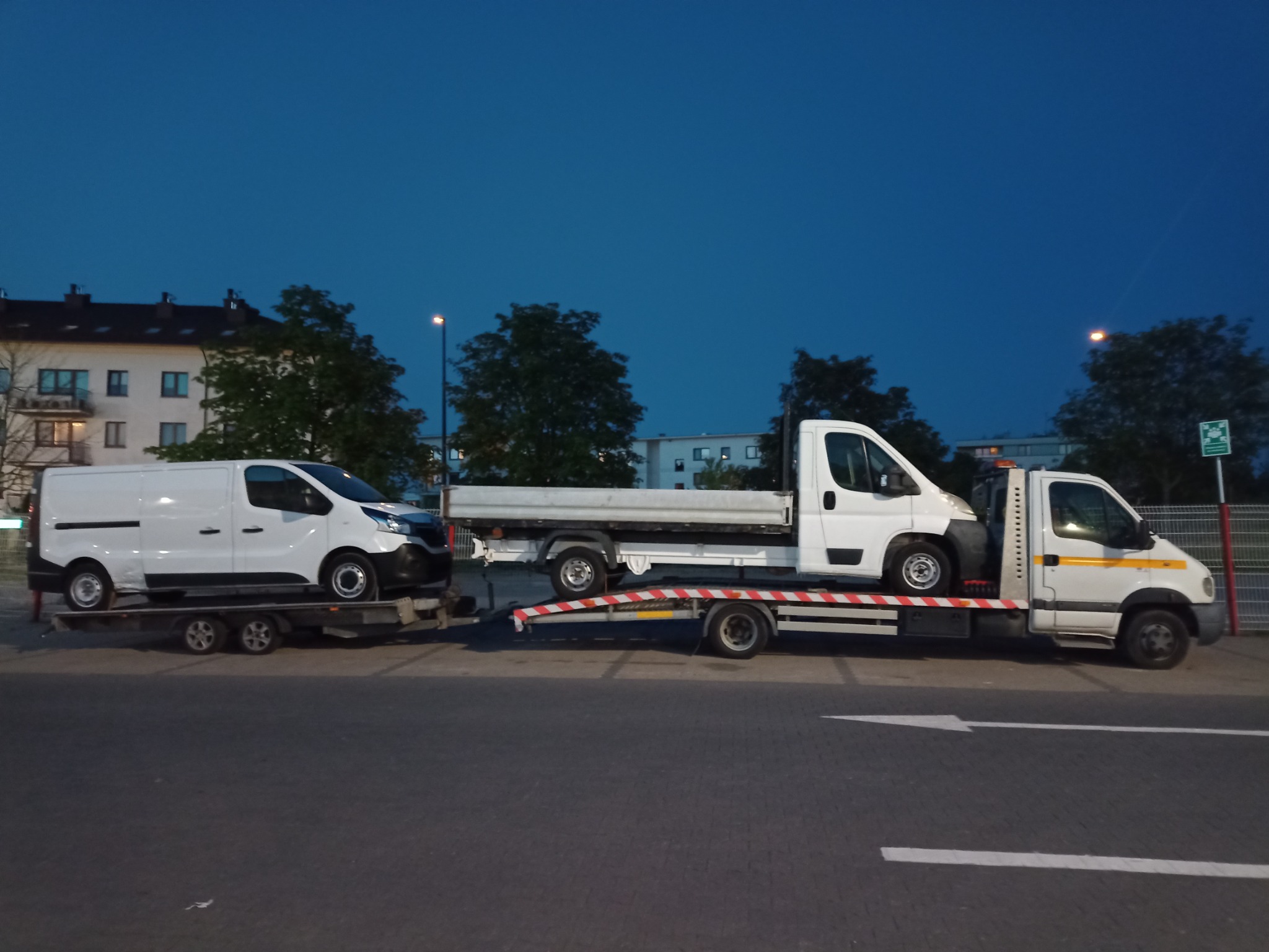 Dwa białe busy transportowane na platformach jednej ciężarówki w miejskim otoczeniu, widoczne budynki mieszkalne i oświetlenie uliczne, ujęcie z poziomu chodnika.