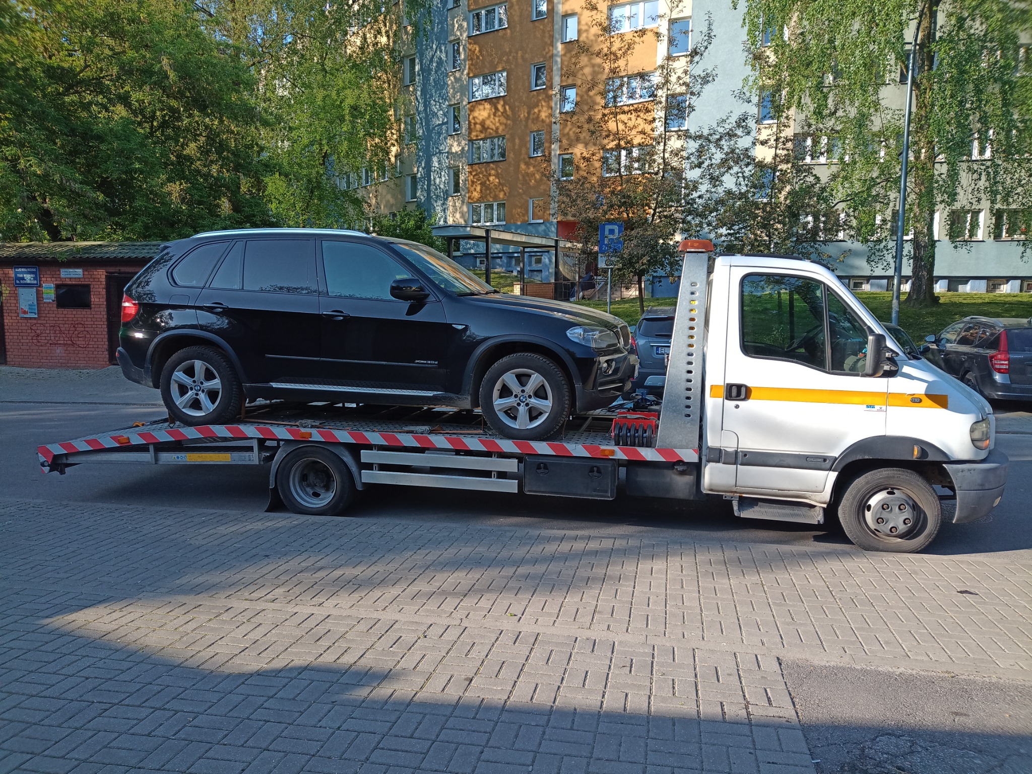 Czarny samochód osobowy marki BMW załadowany na białą platformę pojazdu pomocy drogowej w miejscowości Piaski, na tle bloku mieszkalnego i zieleni.