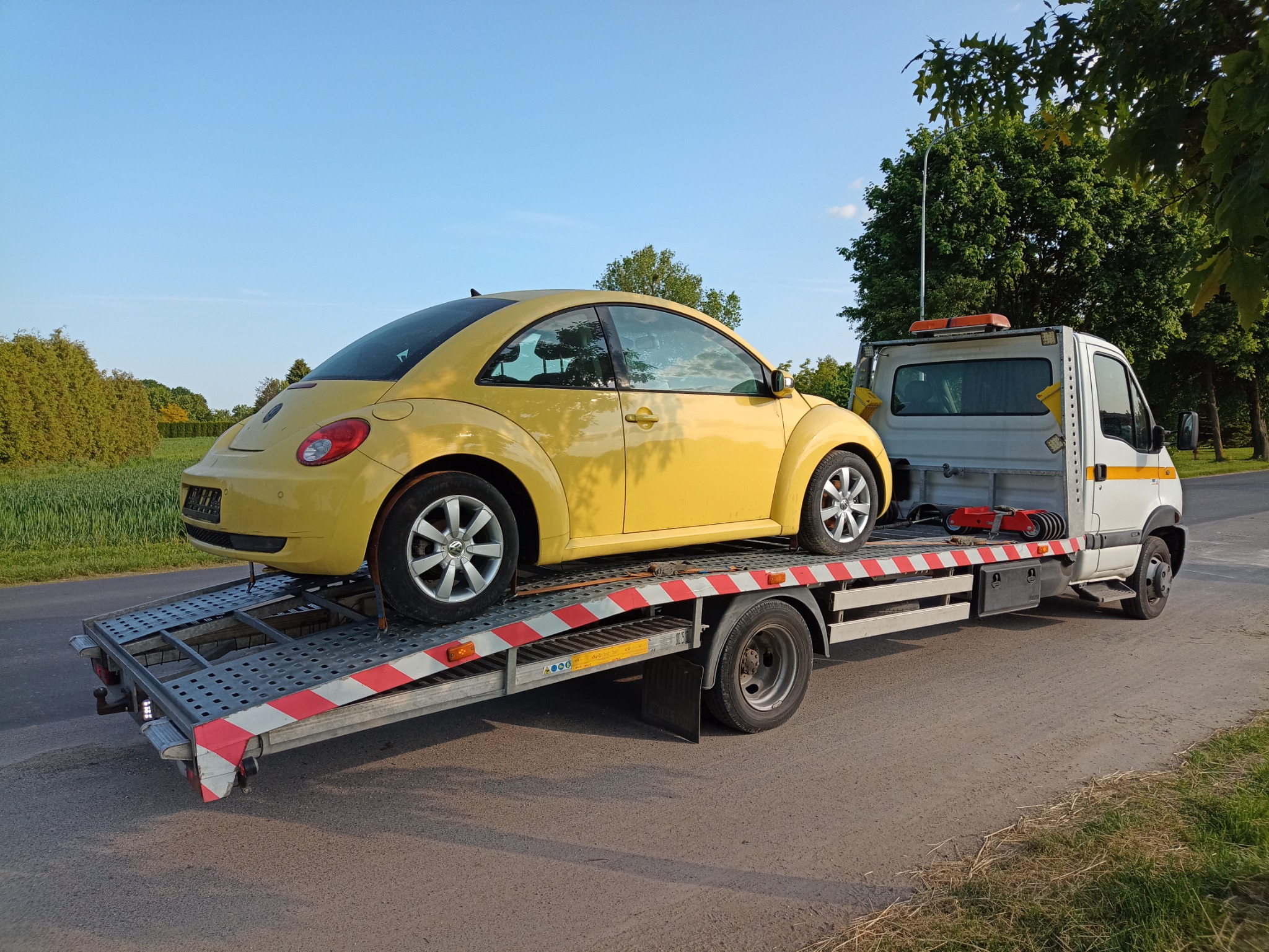 Żółty Volkswagen Beetle transportowany na lawecie w słoneczny dzień, widok z boku, na poboczu drogi.
