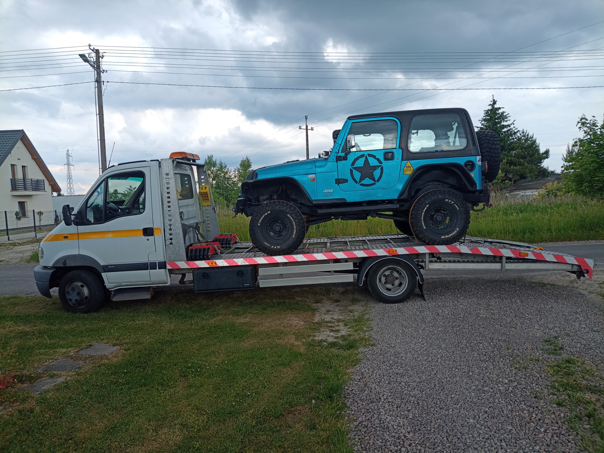 Niebieski Jeep Wrangler z czarną gwiazdą na boku transportowany na platformie pojazdu pomocy drogowej, na tle domu i pochmurnego nieba.